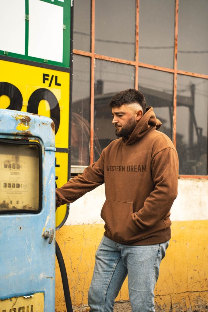 Homme portant un hoodie marron Western Dream devant une ancienne pompe à essence dans un décor western vintage