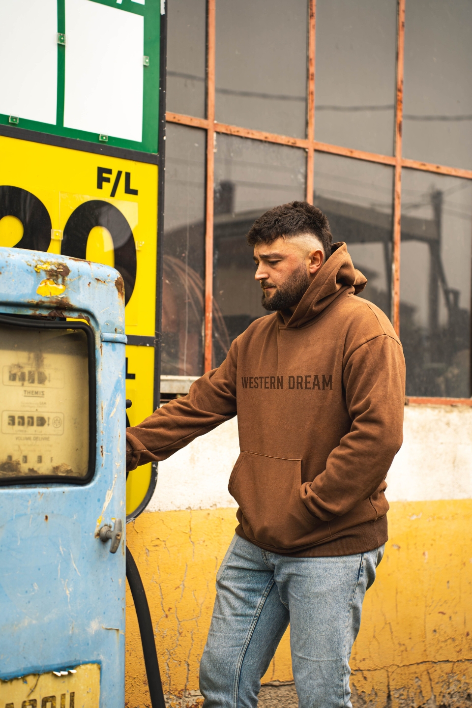 Homme portant un hoodie marron Western Dream devant une ancienne pompe à essence dans un décor western vintage